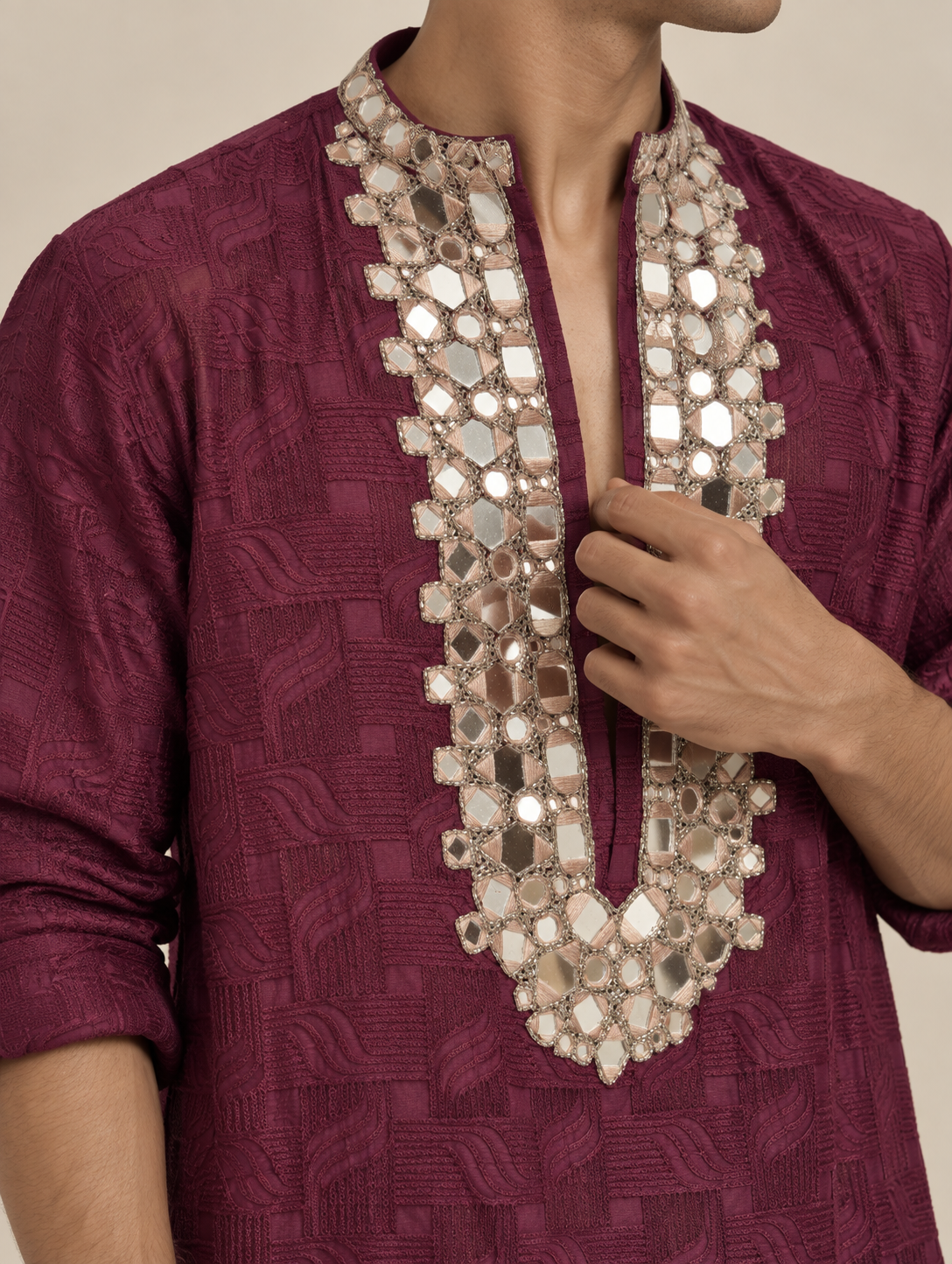A close-up of a person wearing a deep magenta kurta with intricate silver mirror work embroidery around the V-neck collar, with one hand adjusting the collar.